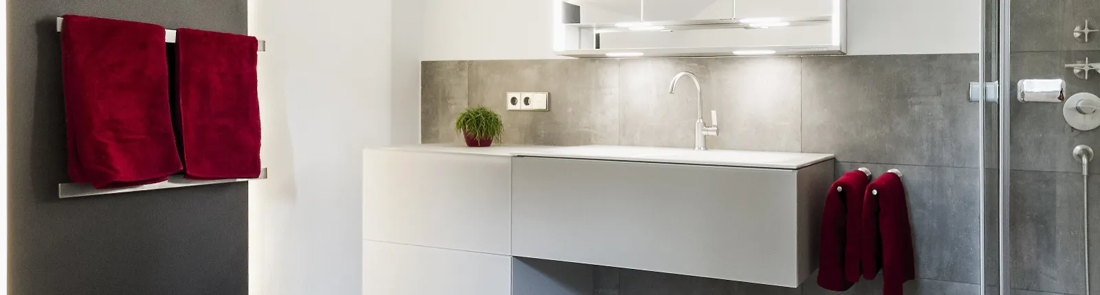 Ein Badezimmer mit grauen Fliesen, einer weißen Waschkonsole und roten Handtüchern. Auf der Konsole steht eine kleine grüne Pflanze, darüber hängt ein beleuchteter Spiegelschrank.