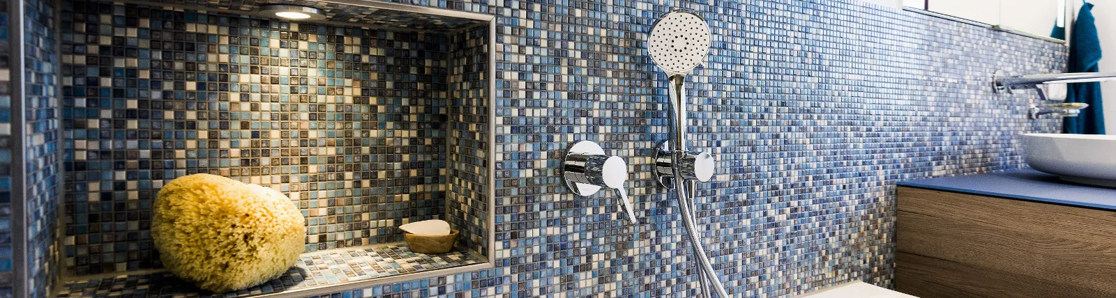 Blau schimmernde Mosaikfliesen an einer Badezimmerwand mit integrierter Nische, die eine Natur-Badeschwamm und Seife zeigt.