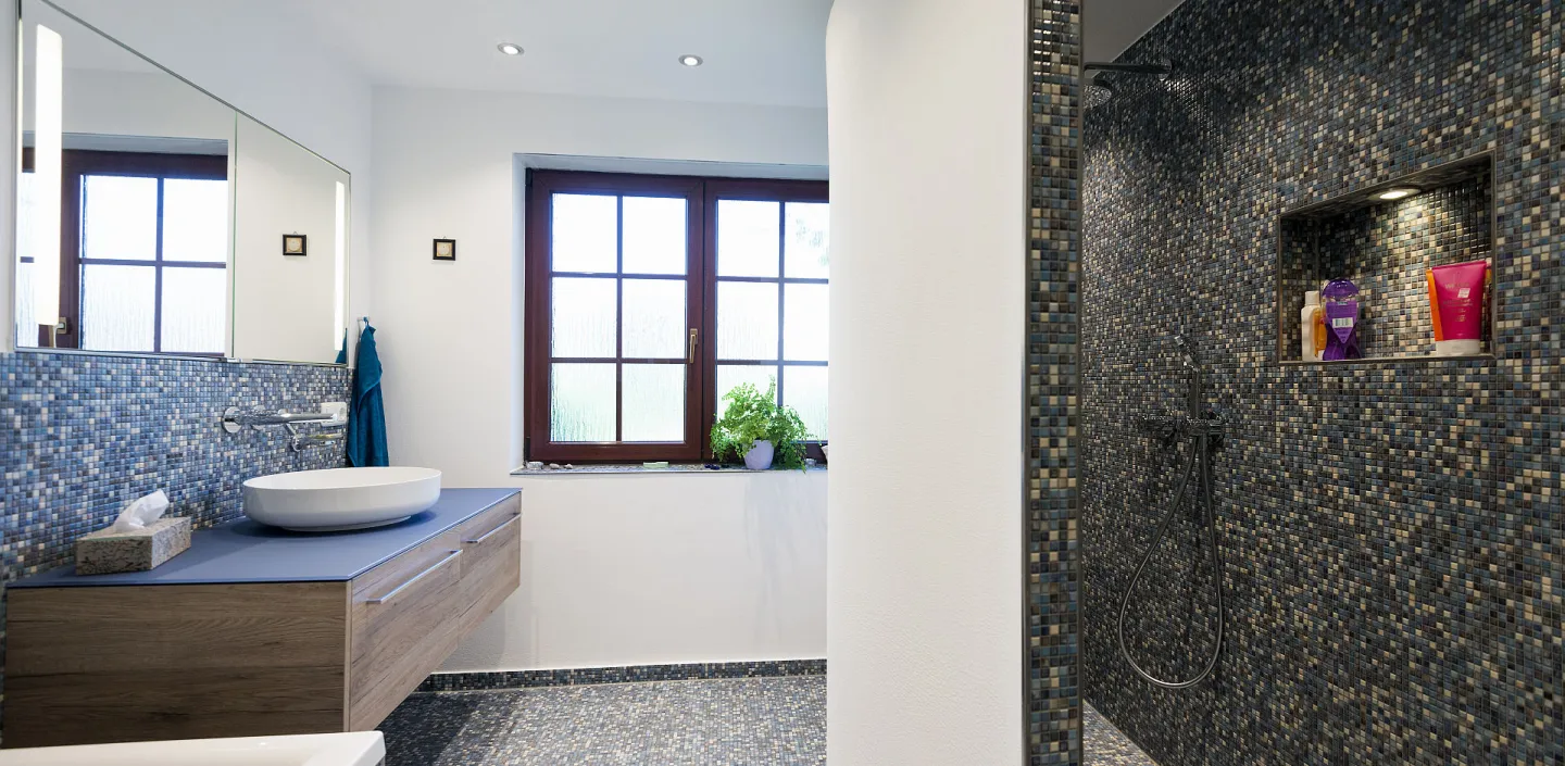 Großes Badezimmer mit blauen Mosaikfliesen, Holzwaschtisch, Glasdusche und großem Fenster.