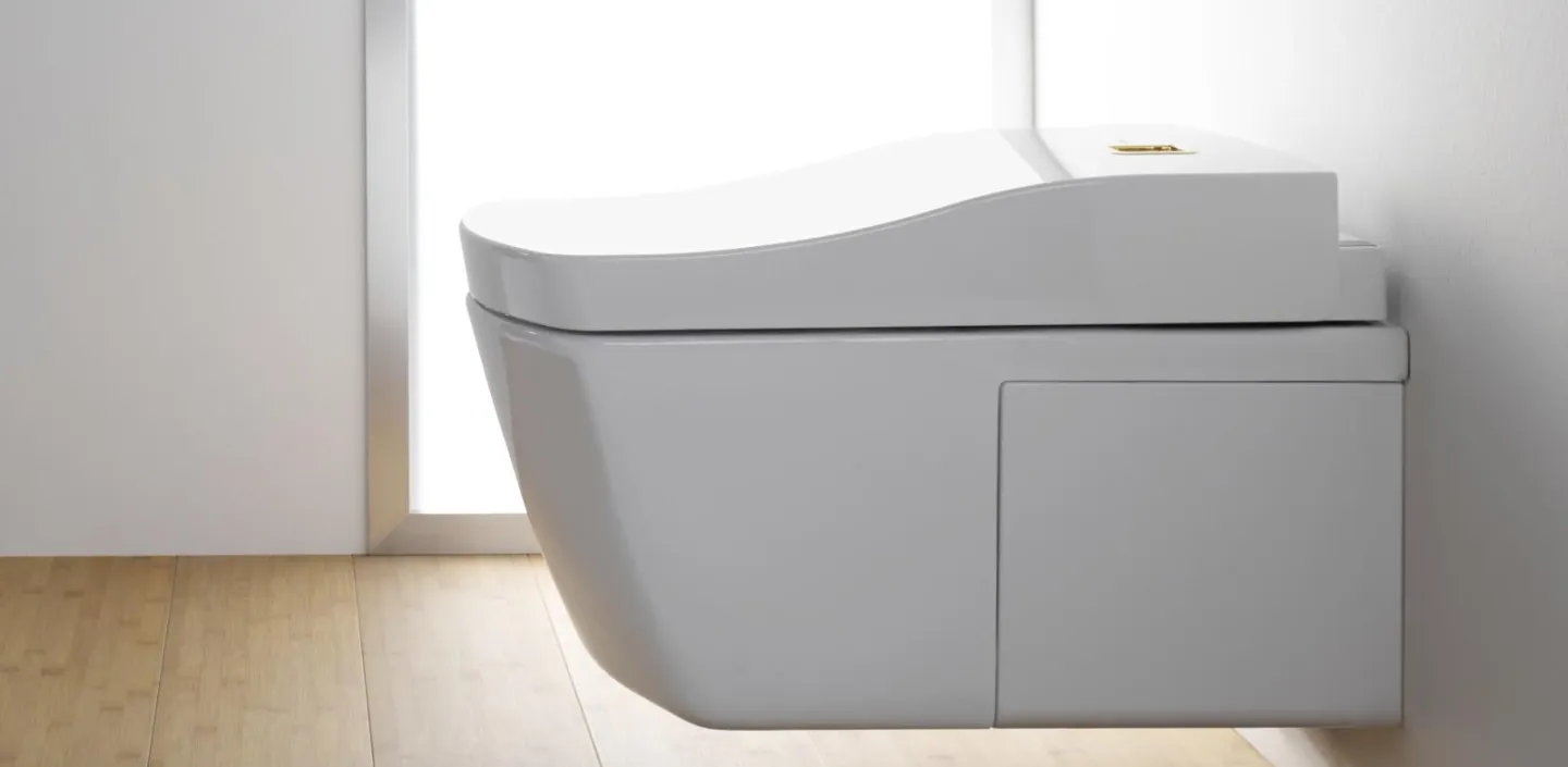 Ein weißes Toto Neorest AC WC, wandhängend und mit abgerundetem Deckel, vor einem großen Fenster und einer weißen Wand, auf Holzfußboden.