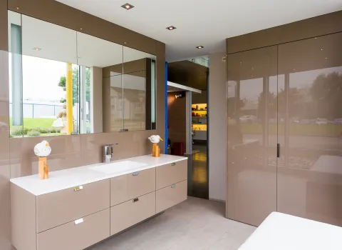 Ein modernes Badezimmer mit glänzenden, beigefarbenen Schränken und einer großen Spiegelfläche, die den Raum optisch erweitert.
