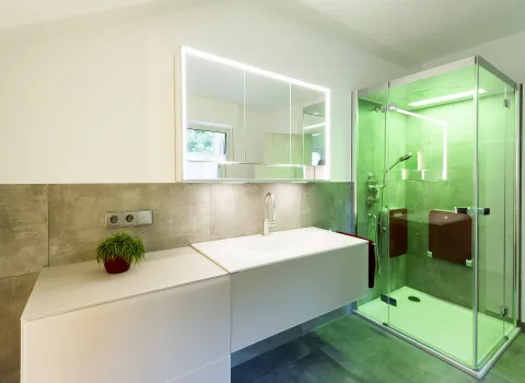 Ein Badezimmer mit einem weißen Waschbereich und einer Glasdusche von Artweger mit grüner Beleuchtung.