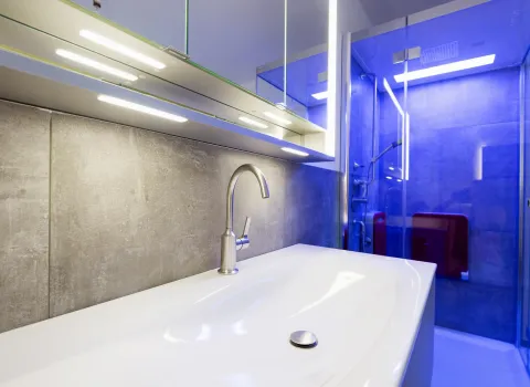 Ein großes Waschbecken mit Chromarmatur von Dornbracht vor einer grauen Fliesenwand. Darüber befinden sich ein beleuchteter Spiegelschrank und LED-Leuchten.