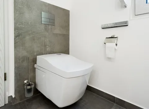 Ein minimalistisches Badezimmer mit einem wandmontierten WASHLET von Toto. Die Rückwand ist mit grauen Fliesen verkleidet, die restlichen Wände sind weiß gestrichen.
