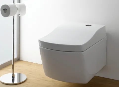 Ein wandhängendes Toto Neorest WC vor einer weißen Wand, auf einem hellen Holzfußboden, daneben ein freistehender Toilettenpapierhalter.