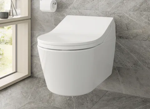 Ein weißes Toto WASHLET RX mit geschlossenem Deckel, vor einer Wand aus grauen Fliesen, daneben ein Fensterbrett mit Vase und eine Wandsteuerung.
