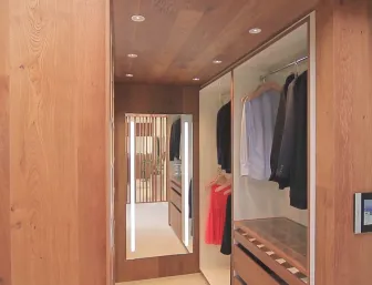 Ein stilvoller, moderner begehbarer Kleiderschrank aus hellem Holz, der mit eingebauten Kleiderstangen, Schubladen und einem beleuchteten Spiegel ausgestattet ist.