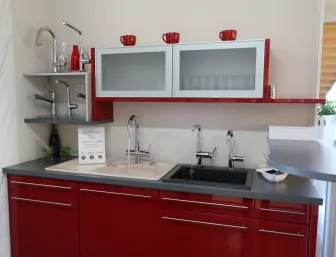 Eine moderne Küche mit glänzend roten Unterschränken und einer grauen Arbeitsplatte, auf der zwei Spülbecken mit jeweils einem Chrom-Wasserhahn installiert sind. 