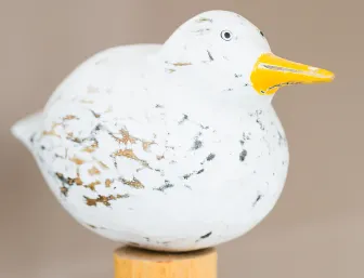 Nahaufnahme einer handgeschnitzten Holzfigur in Form eines Vogels, bemalt in Weiß mit schwarzen und braunen Akzenten, die wie Federn wirken. 