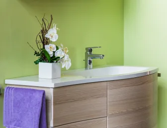 Ein modernes Waschbecken mit abgerundeter, holzähnlicher Kommode steht vor einer hellgrünen Wand.