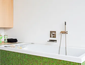 Ein modernes Badezimmer mit einer freistehenden Badewanne, deren Außenseite mit kleinen, grünen Mosaikfliesen verkleidet ist. 