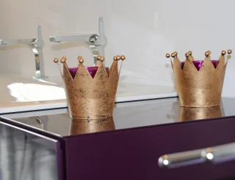 Zwei goldene, kronenförmige Teelichthalter stehen nebeneinander auf einer glänzenden, dunkellila Oberfläche. 