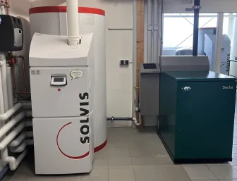 Ein heller Raum mit Fliesenboden zeigt zwei Heizgeräte im direkten Vergleich: links ein Solvis-Heizsystem mit weißem Gehäuse und rotem Rand, rechts das grüne Dachs-Kraftwerk. 