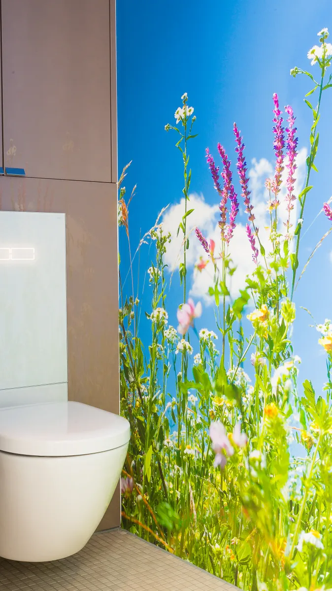 Ein Badezimmer mit einer schwebenden Toilette vor einer großflächigen Wandgestaltung, die eine Wiese mit bunten Wildblumen und einem blauen Himmel zeigt.