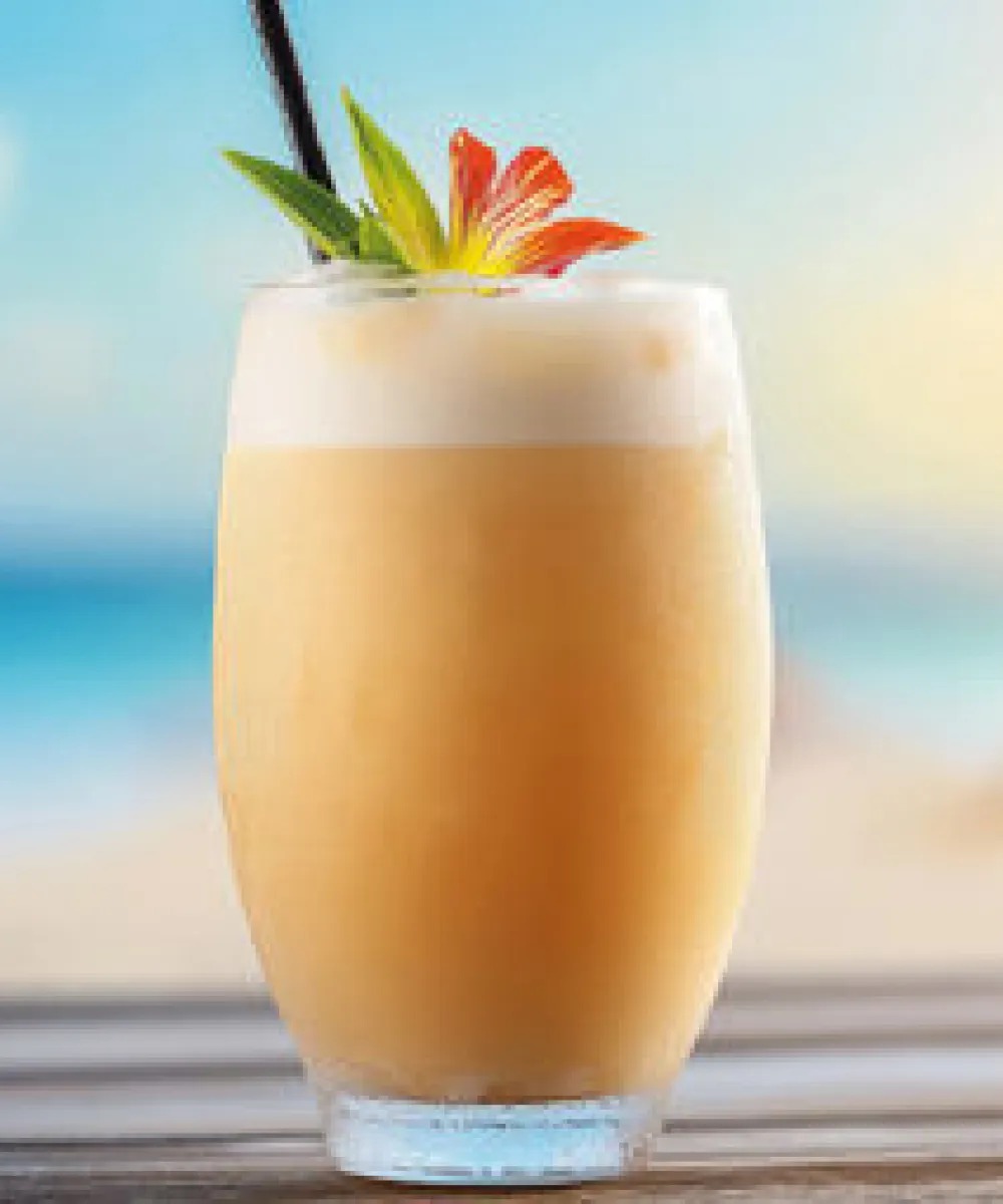 Ein Glas mit einem cremigen Sommer-Cocktail, dekoriert mit einer exotischen Blume und Ananas, steht auf einem Tisch vor einem Strandpanorama.