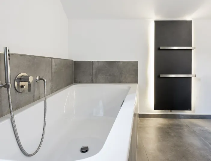 Ein Badezimmer mit einer weißen Badewanne, grauen Fliesen und einem schwarzen Handtuchheizkörper von Bemm.