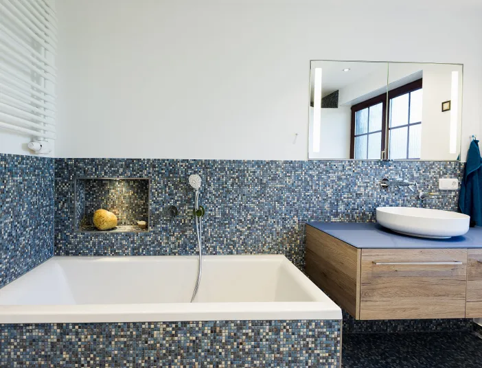 Badezimmer mit blauen Mosaikfliesen, eingebauter Badewanne und beleuchteter Nische. Holzwaschtisch mit Aufsatzbecken und großem Spiegel.