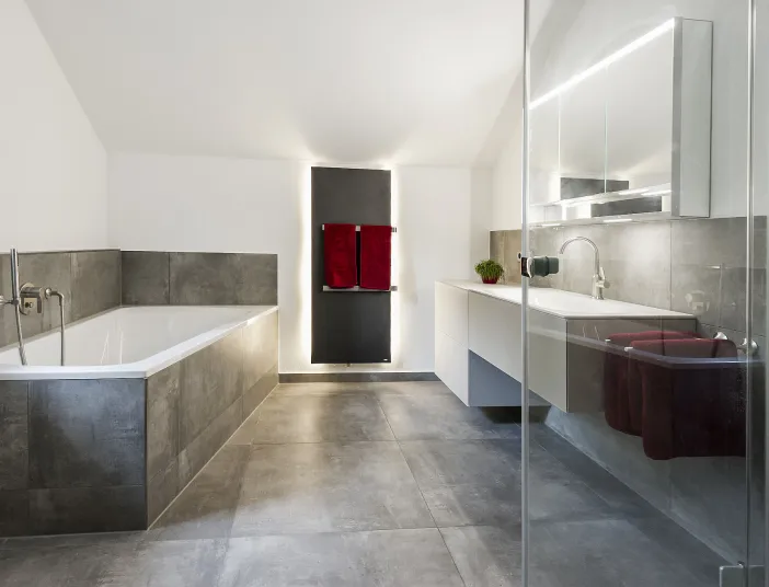 Ein geräumiges Badezimmer mit grauer Fliesenverkleidung, einer Badewanne auf der linken Seite und einem beleuchteten Waschtisch rechts. 