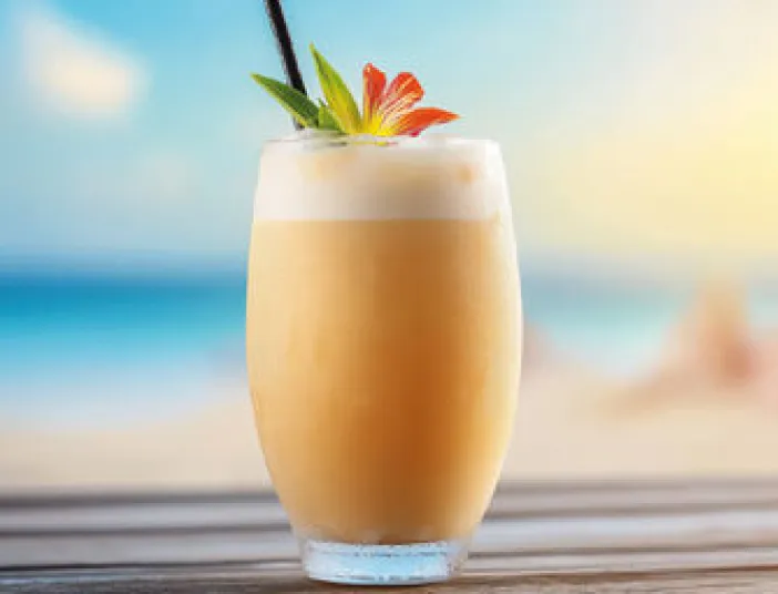 Ein Glas mit einem cremigen Sommer-Cocktail, dekoriert mit einer exotischen Blume und Ananas, steht auf einem Tisch vor einem Strandpanorama.